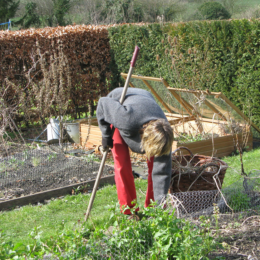 Eine Person in einem grauen Pullover und einer roten Hose, die mit einer Hacke im Garten Frühjahrsarbeiten verrichtet, inspiriert von Sa., 13.3.2027 | 11.00 – 12.30 Uhr GARTENTALK – Gedanken zum Gärtnern im Frühling.