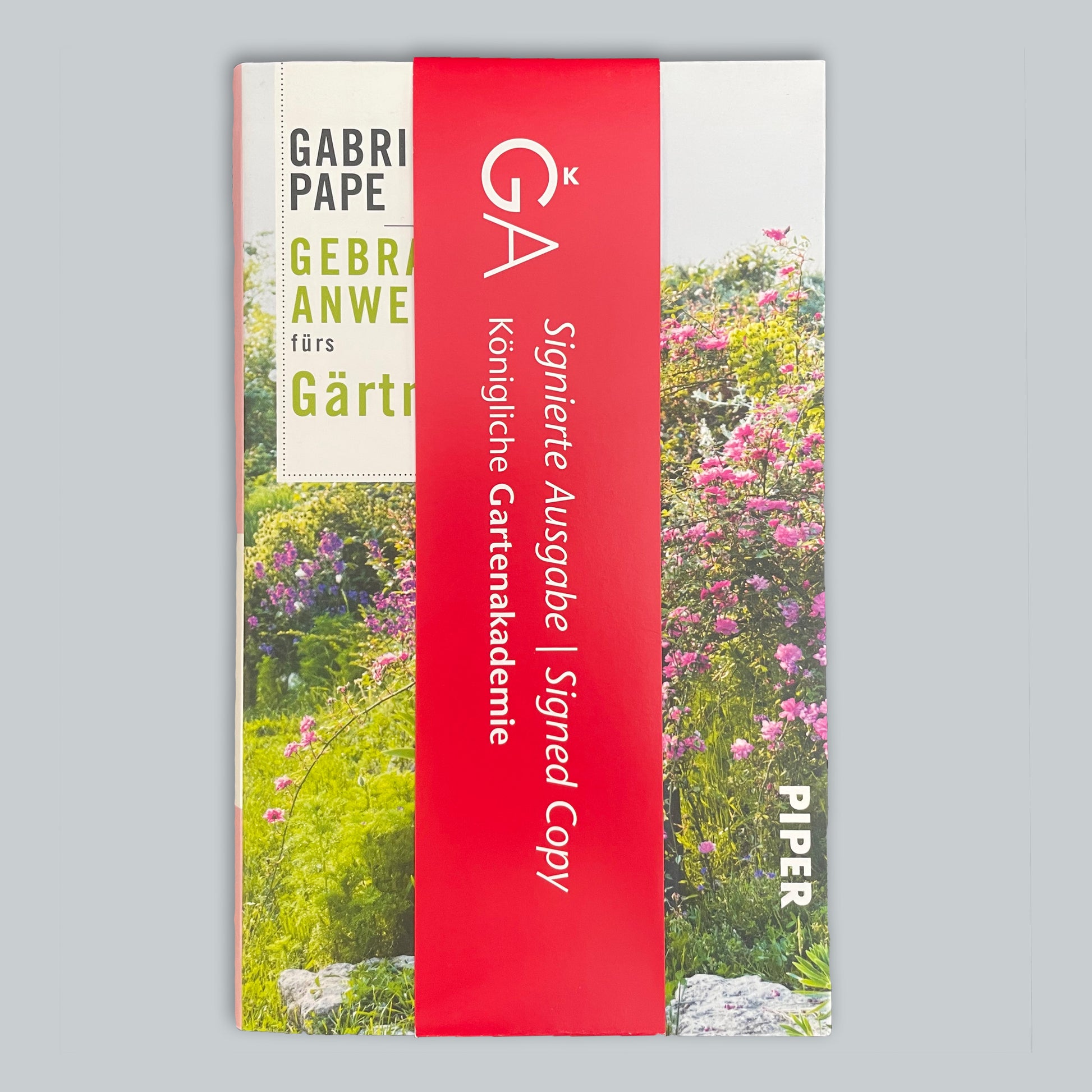 Gebrauchsanweisung fürs Gärtnern | Praxis & Tipps Buch von Gabriella Pape ist teilweise in ein rotes Band mit der Aufschrift "GA Signierte Ausgabe | Signed Copy Königliche Gartenakademie" eingewickelt. Der Einband zeigt eine lebendige Gartenszene.