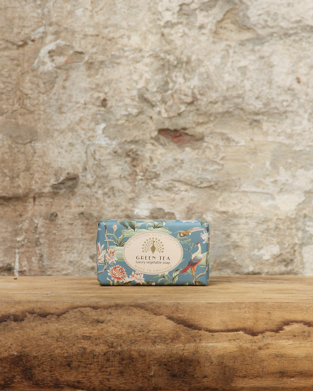 Eine Tafel ESC | 'Green Tea' frisch duftende pflanzliche Handseife von The English Soap Company in blumiger, plastikfreier Verpackung liegt auf Holz vor einer hellen Steinwand.