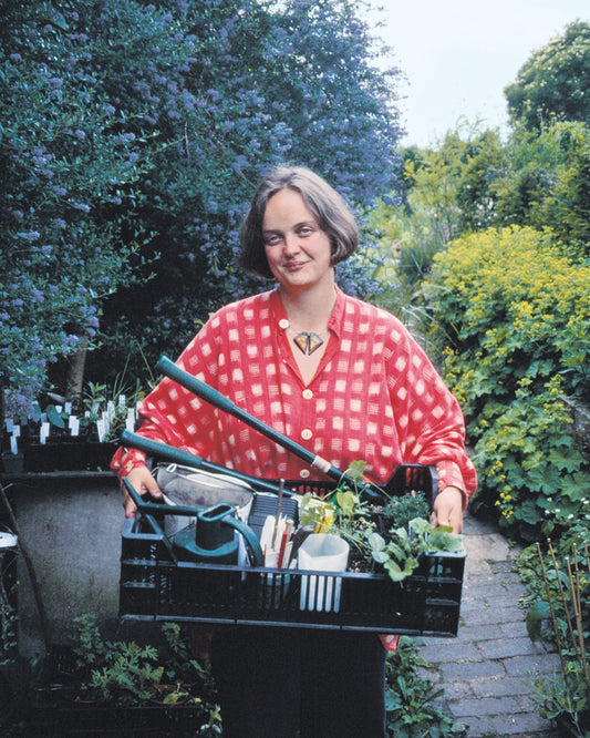 Beim HALBTAGESKURS der Königlichen Gartenakademie "Der Cottage Garten: Der Garten der Zukunft" (07.03.2026, 13:30-16:30) steht eine Person mit einer Kiste voller Werkzeuge und Pflanzen auf dem Gartenweg, umgeben von üppigem Grün der Gartenschule.