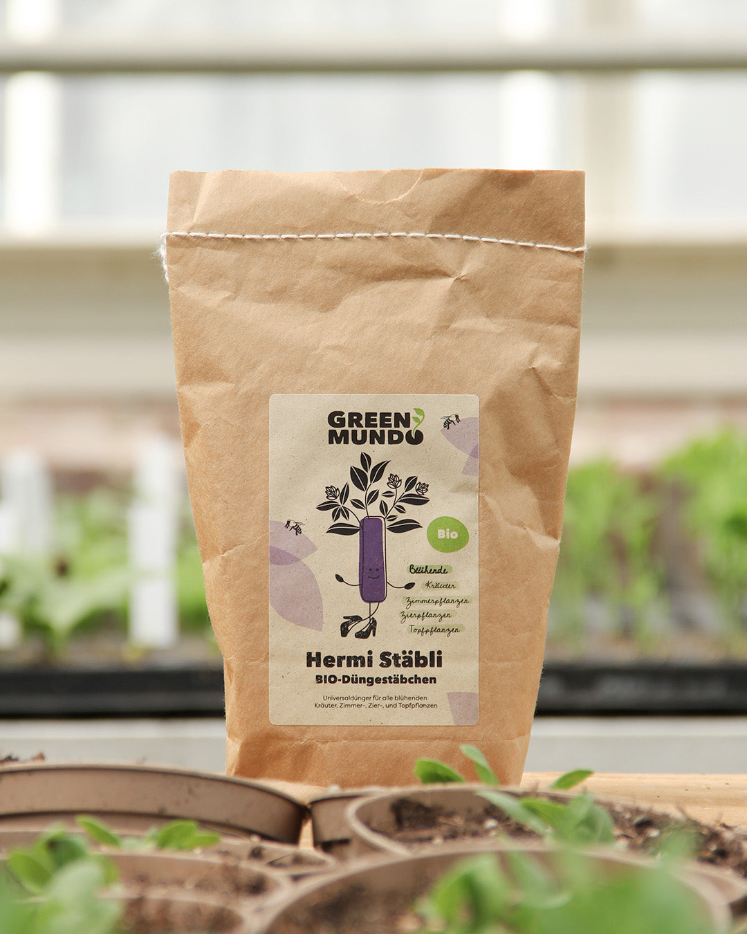 Eine braune Papiertüte mit der Aufschrift Green Mundo | 'Hermi' Bio-Universaldüngestäbchen 300 g steht auf einer Holzfläche mit kleinen Grünpflanzen und Töpfen im Hintergrund. Auf dem Etikett sind Blätter und ein lila Stab abgebildet.