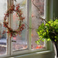 Ein sonnendurchflutetes Fenster mit einer roten Beerenkränze, einer Blattpflanze und Herbstblättern stimmt auf den "Di. 29.09.2026 | 11.00-12.30 WORKSHOP Herbstliches Türkranzbinden" der Königlichen Gartenakademie - die perfekte Inspiration für Ihre nächste Bastelstunde!.