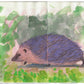 Aquarell-Illustration eines Igels mit braunem Gesicht und violetten Stacheln auf Gras, gemalt auf zwei Skizzenbuchseiten während des Königlichen Gartenakademie AQUARELL WORKSHOP Tiere Skizzieren NEU (Fr. 02.10.2026 | 10:00-13:00).