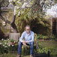 Ein Mann in Jeans und hellblauem Hemd sitzt in einem üppigen Garten, Gartengeräte und Topfpflanzen in der Nähe, bereit für den "HALBTAGESKURS Das Gartenjahr-Monat für Monat mit Horst Mager" in der Königlichen Gartenakademie am Fr. 15.05.2026, 13:00-16:00.