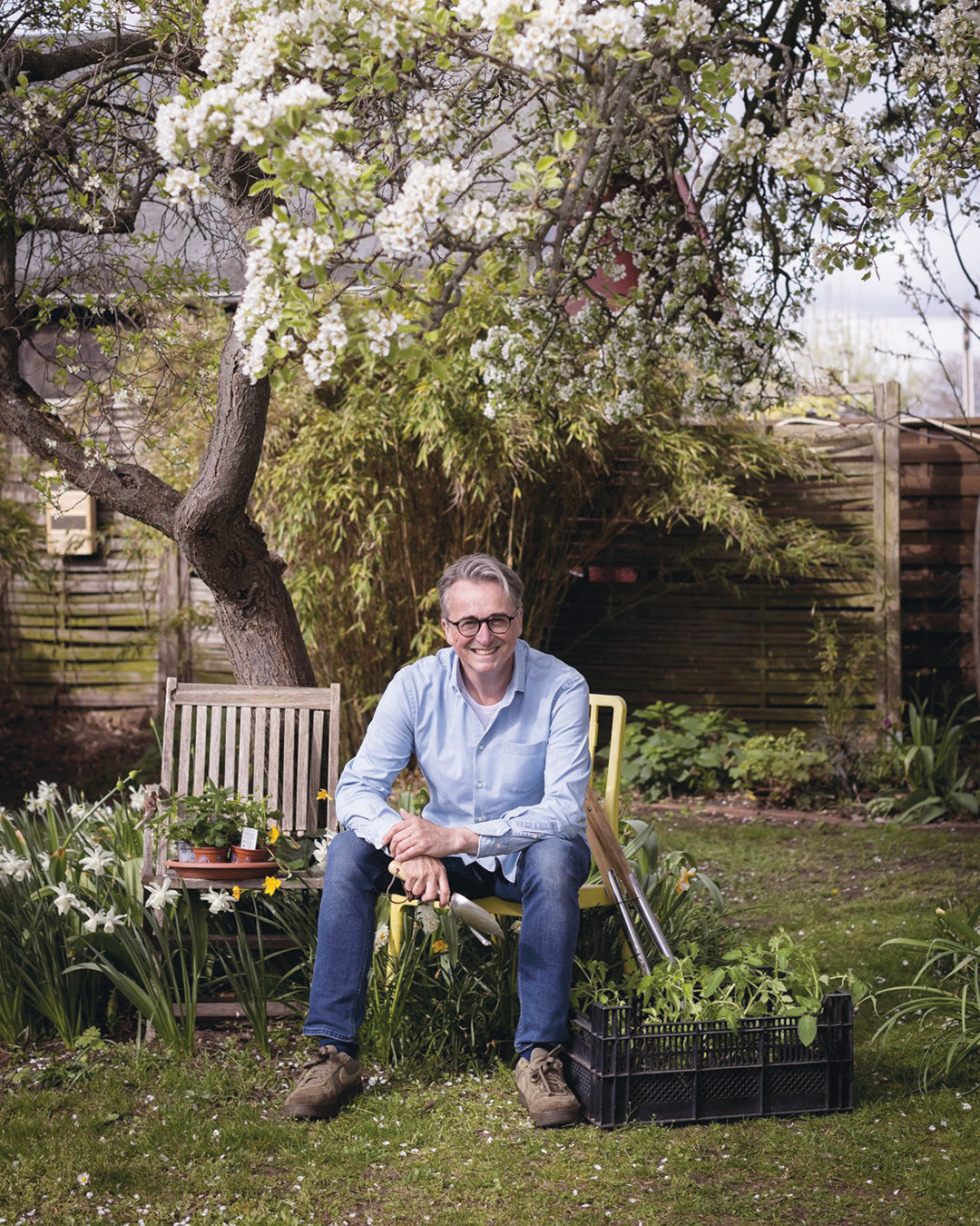Ein Mann in Jeans und hellblauem Hemd sitzt in einem üppigen Garten, Gartengeräte und Topfpflanzen in der Nähe, bereit für den "HALBTAGESKURS Das Gartenjahr-Monat für Monat mit Horst Mager" in der Königlichen Gartenakademie am Fr. 15.05.2026, 13:00-16:00.