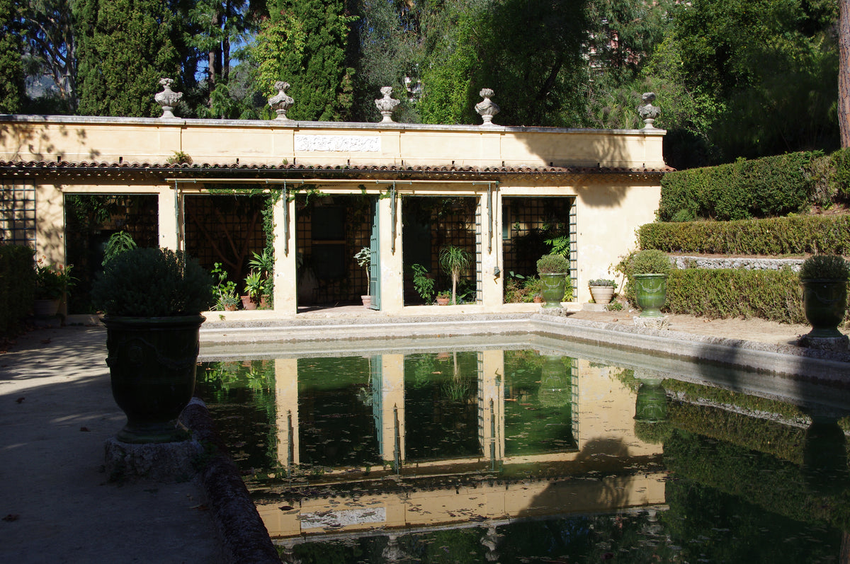 Ein rechteckiger Gartenpool spiegelt einen gelben Steinpavillon wider. Am 16.3.2027 | 14.30 – 15.30 Uhr AFTERNOON TEA Gartenreise durch Frankreich, mit offenen Türen und Steinverzierungen entlang der Dachlinie.