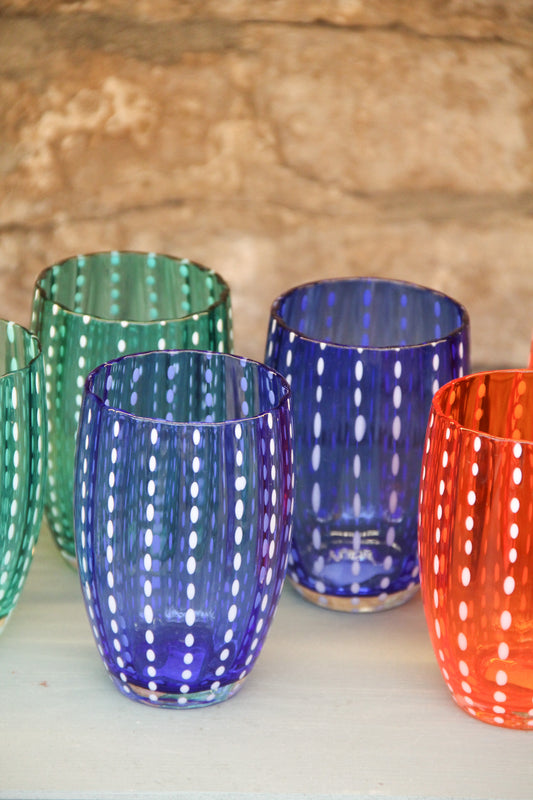 Vier farbenfrohe Zafferano | Perle-Gläser in verschiedenen Farben (grün, blau und orange, jeweils 32 cl) mit vertikalen Linien aus weißen Punkten sind auf einer hellen Fläche vor einem strukturierten Stein-Hintergrund angeordnet. Diese mundgeblasenen Gläser von Zafferano stehen für feine italienische Glaskunst.