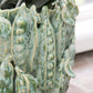 Eine Nahaufnahme der Vase „Vase grün mit Schoten“ (H 16, Ø 18 cm) von Des Pots mit erhabenen Erbsenschoten-Akzenten und einer grünen Pflanze darin ist ein bezauberndes Deko-Highlight, wenn sie in Fensternähe platziert wird.