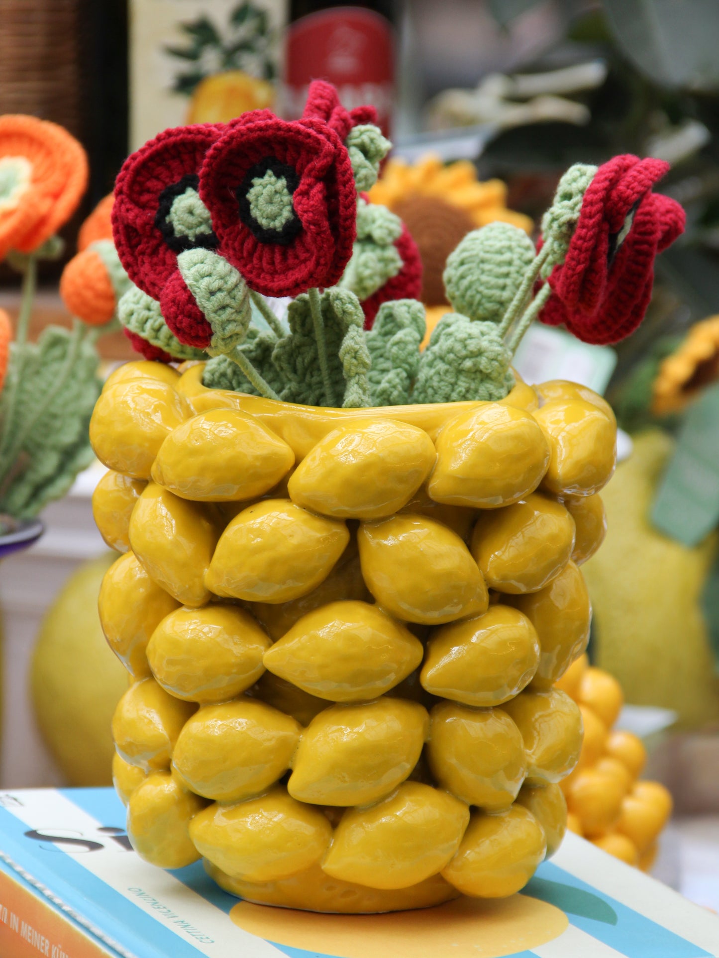 Ein schmaler Blumentopf „Des Pots“ mit Zitronenmotiv in Gelb (H 16 Ø 18 cm) von Des Pots beherbergt rote und grüne gestrickte Blumen. Der Keramiktopf verkörpert mediterrane Eleganz und steht auf einem Buch, während im Hintergrund gelbe und orangefarbene Dekorationselemente unscharf zu erkennen sind.