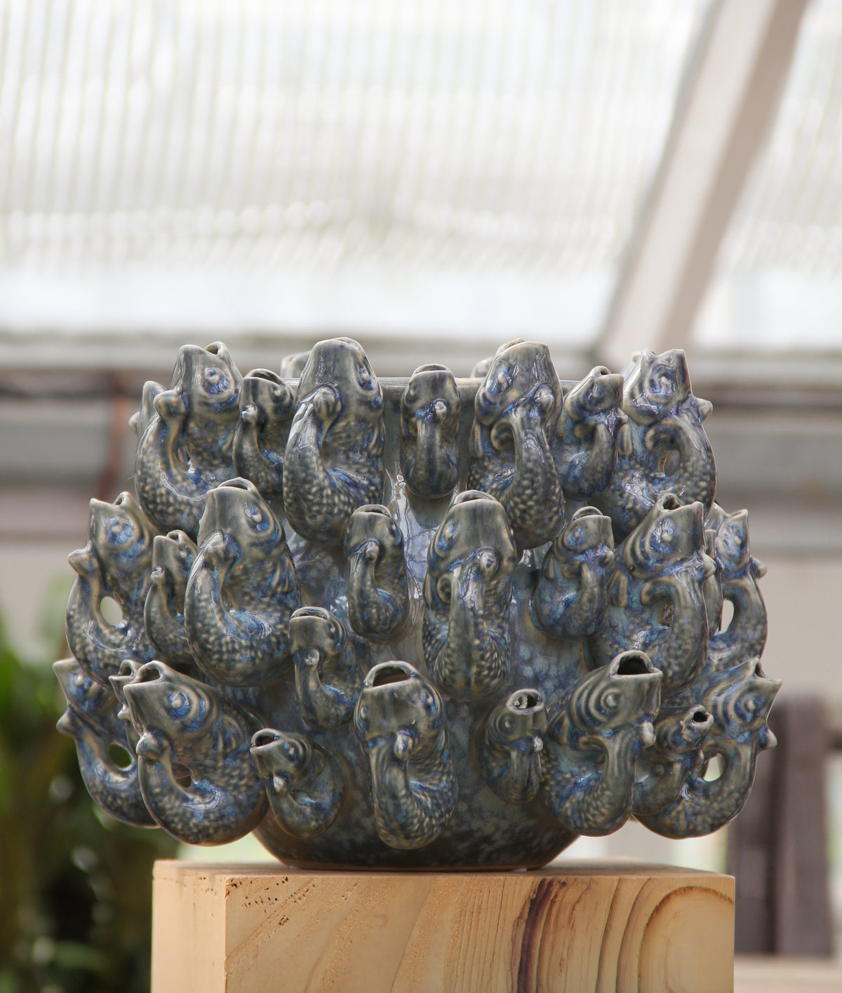 The Des Pots | Vase „Fisch“ klein in Blau aus Steingut (H 18 cm, Ø 23 cm) von Des Pots, eine blaue Keramikvase aus Steingut in Form eines Fischschwarms mit geöffneten Mäulern, wird im Innenraum auf einem Holzsockel vor dem Hintergrund eines verschwommenen Gewächshauses präsentiert.