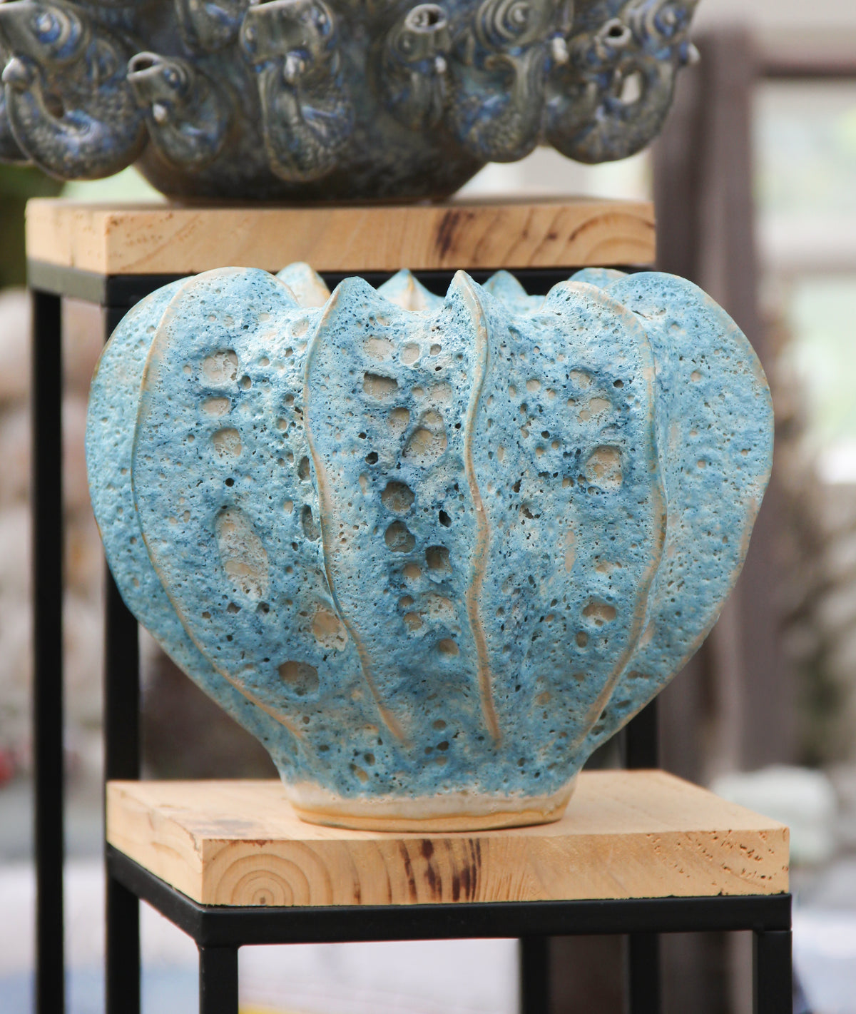 Eine blaugrüne Vase „Moos“ aus Steingut (H 16, Ø 20 cm) von Des Pots mit vertikalen Rillen und dekorativen Öffnungen steht auf einem hellen Holzständer. Diese wasserdichte Keramikvase hat eine strukturierte Oberfläche, während im Hintergrund ein weiteres Keramikstück sanft verschwommen erscheint.
