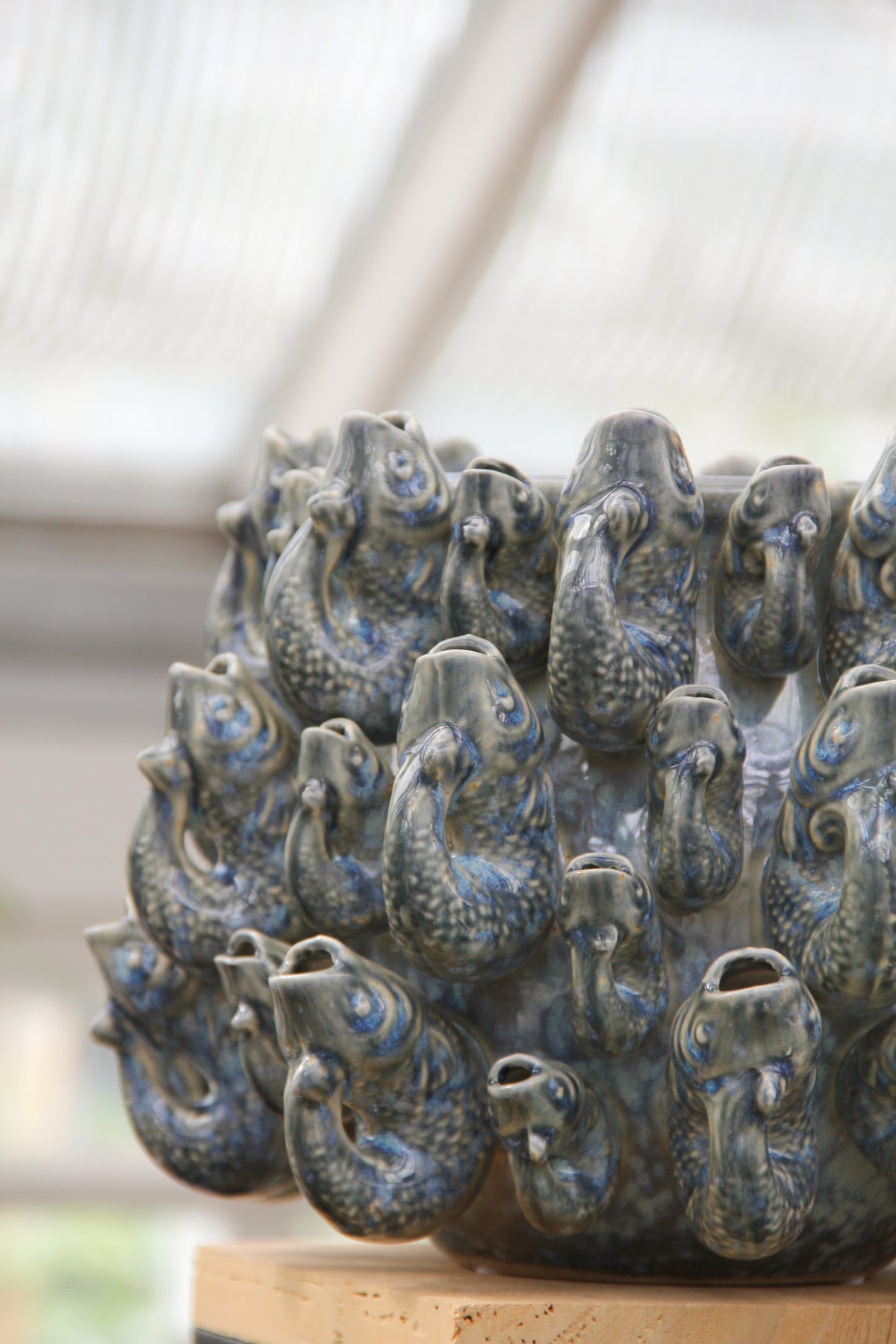 Wir stellen vor: Des Pots | Vase „Fisch“, klein, in Blau aus Steingut (H 18 cm, Ø 23 cm) von Des Pots – ein handgeformtes Designstück aus Steingut, glasiert in Blautönen, das mit mehreren aneinandergereihten Fischköpfen und geöffneten Mündern beeindruckt. Diese außergewöhnliche Designer-Vase verbindet kunstvolle Handwerkskunst mit markanter optischer Ausstrahlung und wird stilvoll auf einem Holzsockel im Innenbereich präsentiert.