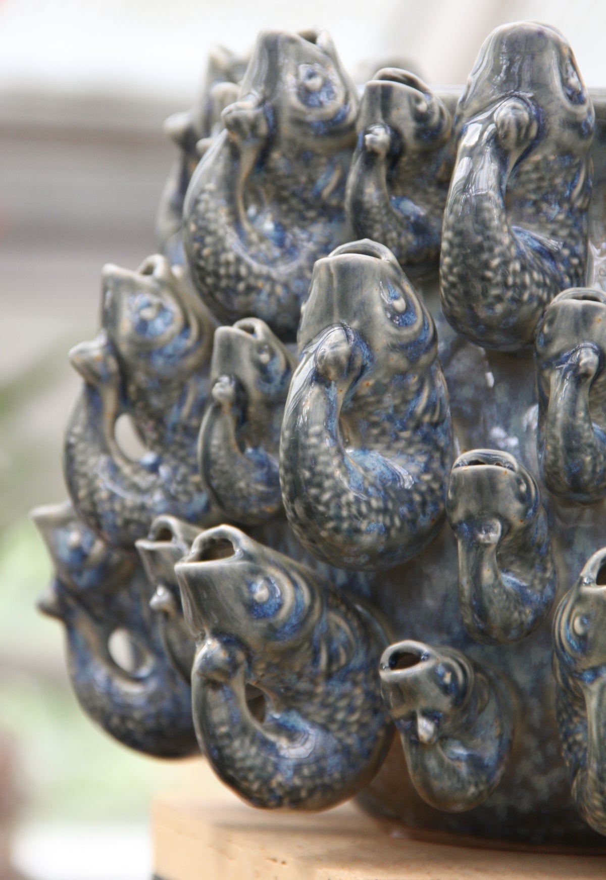 Die Vase „Fisch“ klein in Blau aus Steingut (H 18 cm, Ø 23 cm) von Des Pots ist ein markantes Keramik-Dekorationsstück, das aus mehreren handgeformten Steingutfischen mit geöffneten Mäulern besteht, die kreisförmig um die zentrale Vase angeordnet sind. Jeder Fisch ist mit einer glänzenden blaugrauen Glasur versehen und weist eine detailreiche Schuppenstruktur auf.