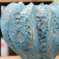 Eine Nahaufnahme der Des-Pots-Vase „Moos“ in Blaugrün aus Steingut (H 16, Ø 20 cm), handgeformt mit unregelmäßigen Löchern und vertikalen Rillen, glasiert und auf einem Holzregal ausgestellt.