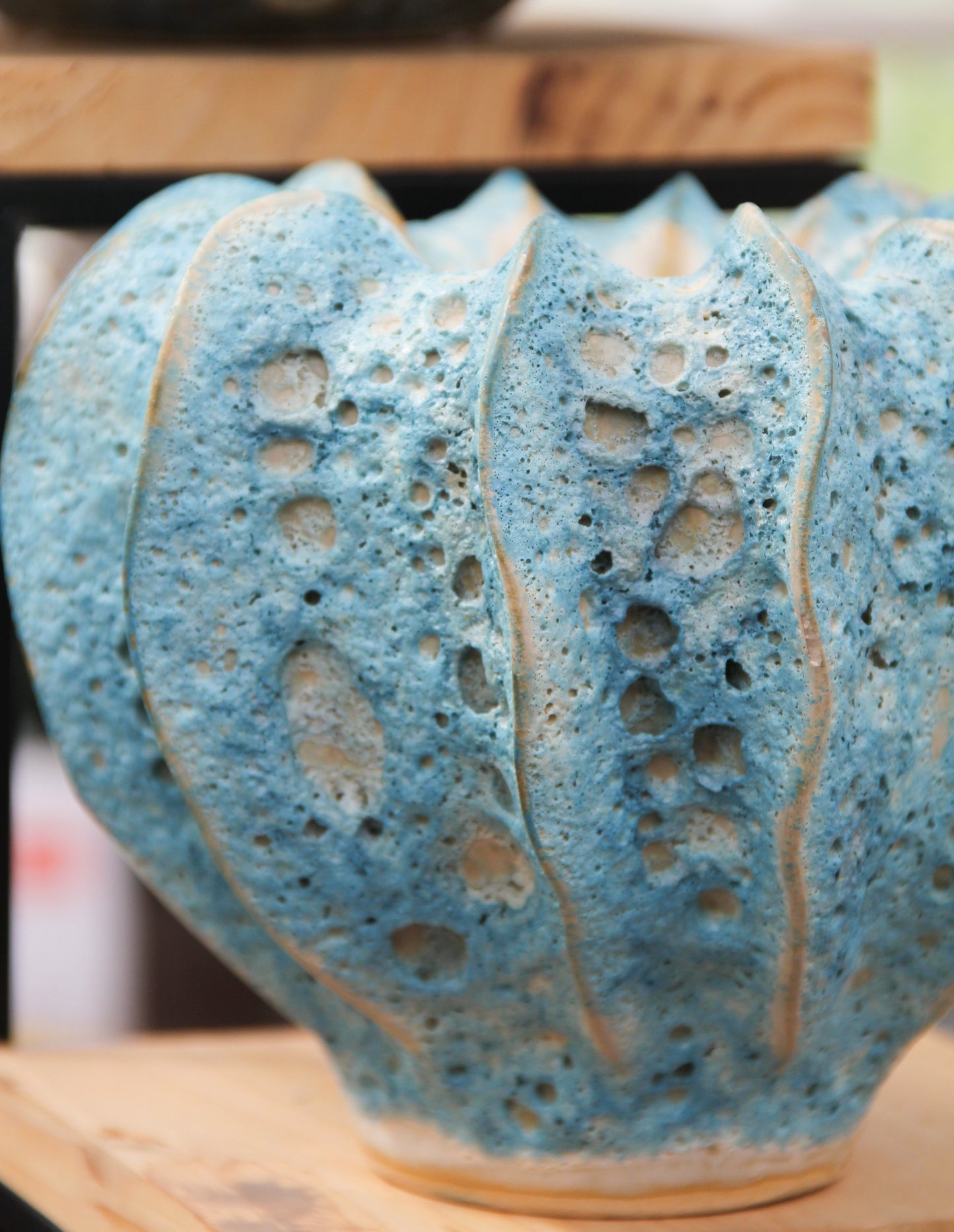 Eine Nahaufnahme der Des-Pots-Vase „Moos“ in Blaugrün aus Steingut (H 16, Ø 20 cm), handgeformt mit unregelmäßigen Löchern und vertikalen Rillen, glasiert und auf einem Holzregal ausgestellt.