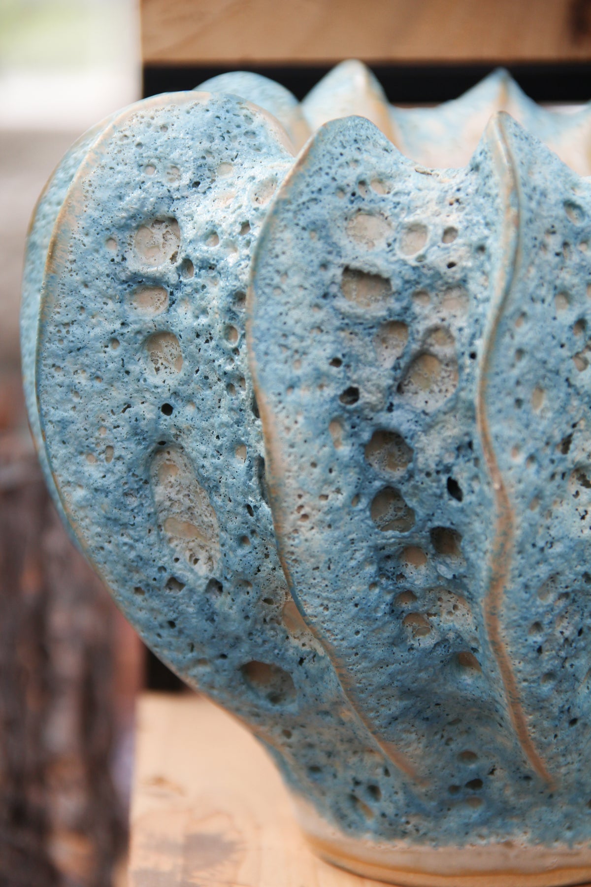 Nahaufnahme der Vase „Moos“ von Des Pots | Blaugrüne Vase aus Steingut, H 16, Ø 20 cm, mit blaugrüner Glasur, wellenförmigen Rippen und porösen, kraterartigen Löchern. Diese handgeformte Steingutvase ist auf einer Holzunterlage ausgestellt.