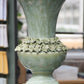 Die „Amphore in Grün aus Steingut, 25 x 41,5 cm“ von Werner Voß, mit ausgestellter Kante und einem Keramik-Blumenring in der Mitte, steht im Innenraum in der Nähe von Regalen und Büchern und verleiht dem Raum einen Hauch mediterraner Dekoration.