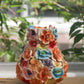 Die Des Pots | Vase mit bunten Deko-Blumen (H 17, Ø 5,5–14 cm) der Marke Des Pots, handgefertigt und mit farbenfrohen, detailreichen Blumenskulpturen verziert, steht auf einer Holzunterlage. Im Hintergrund sorgen grüne Pflanzen und einfallendes Sonnenlicht für eine stilvolle Atmosphäre – das perfekte Wohnaccessoire für jeden Raum.