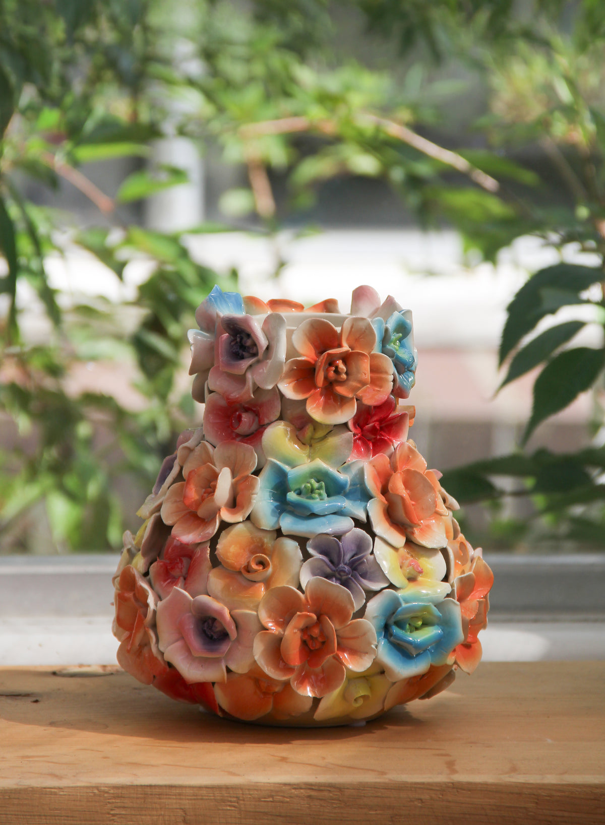Die Des Pots | Vase mit bunten Deko-Blumen (H 17, Ø 5,5–14 cm) der Marke Des Pots, handgefertigt und mit farbenfrohen, detailreichen Blumenskulpturen verziert, steht auf einer Holzunterlage. Im Hintergrund sorgen grüne Pflanzen und einfallendes Sonnenlicht für eine stilvolle Atmosphäre – das perfekte Wohnaccessoire für jeden Raum.
