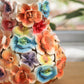 Die „Des Pots | Vase mit bunten Deko-Blumen, H 17, Ø 5,5–14 cm“ von Des Pots ist eine handgefertigte Keramikvase, verziert mit leuchtenden, glänzenden 3D-Blumen in Orange, Blau, Gelb, Lila und Pink. Sie steht auf einer Holzunterlage vor einem verschwommenen grünen Hintergrund.