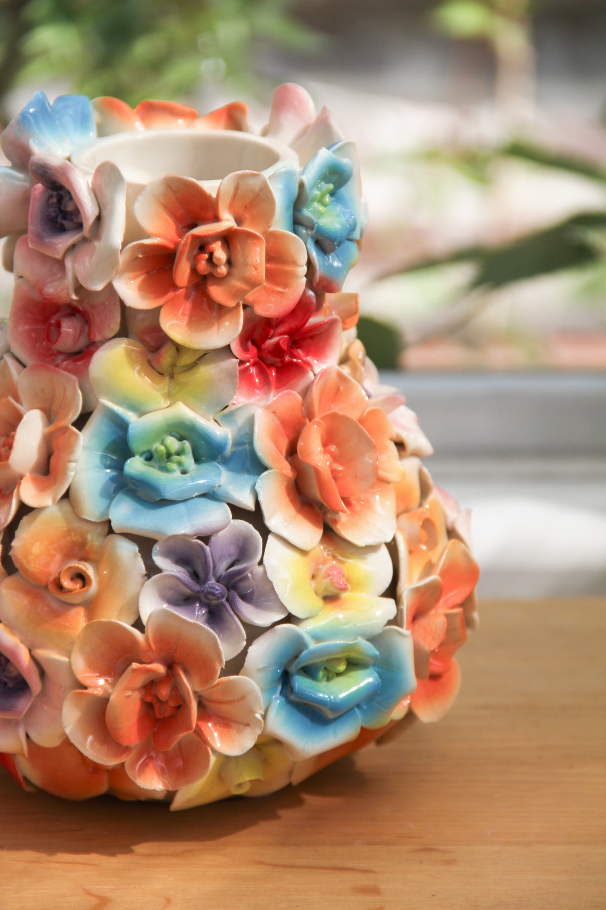 Die „Des Pots | Vase mit bunten Deko-Blumen, H 17, Ø 5,5–14 cm“ von Des Pots ist eine handgefertigte Keramikvase, verziert mit leuchtenden, glänzenden 3D-Blumen in Orange, Blau, Gelb, Lila und Pink. Sie steht auf einer Holzunterlage vor einem verschwommenen grünen Hintergrund.