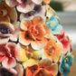 Eine Nahaufnahme von leuchtenden, bunten Keramikblumen mit glänzenden Blütenblättern, kunstvoll arrangiert in der „Des Pots | Vase mit Deko-Blumen bunt“ von Des Pots, die Farbtöne von Orange, Gelb, Rosa, Violett und Blau in der handgefertigten Vase (H 17, Ø 5,5–14 cm).