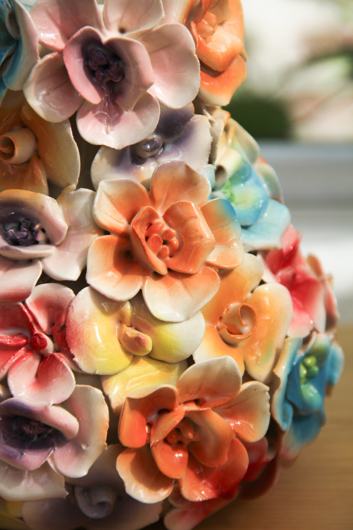 Eine Nahaufnahme von leuchtenden, bunten Keramikblumen mit glänzenden Blütenblättern, kunstvoll arrangiert in der „Des Pots | Vase mit Deko-Blumen bunt“ von Des Pots, die Farbtöne von Orange, Gelb, Rosa, Violett und Blau in der handgefertigten Vase (H 17, Ø 5,5–14 cm).