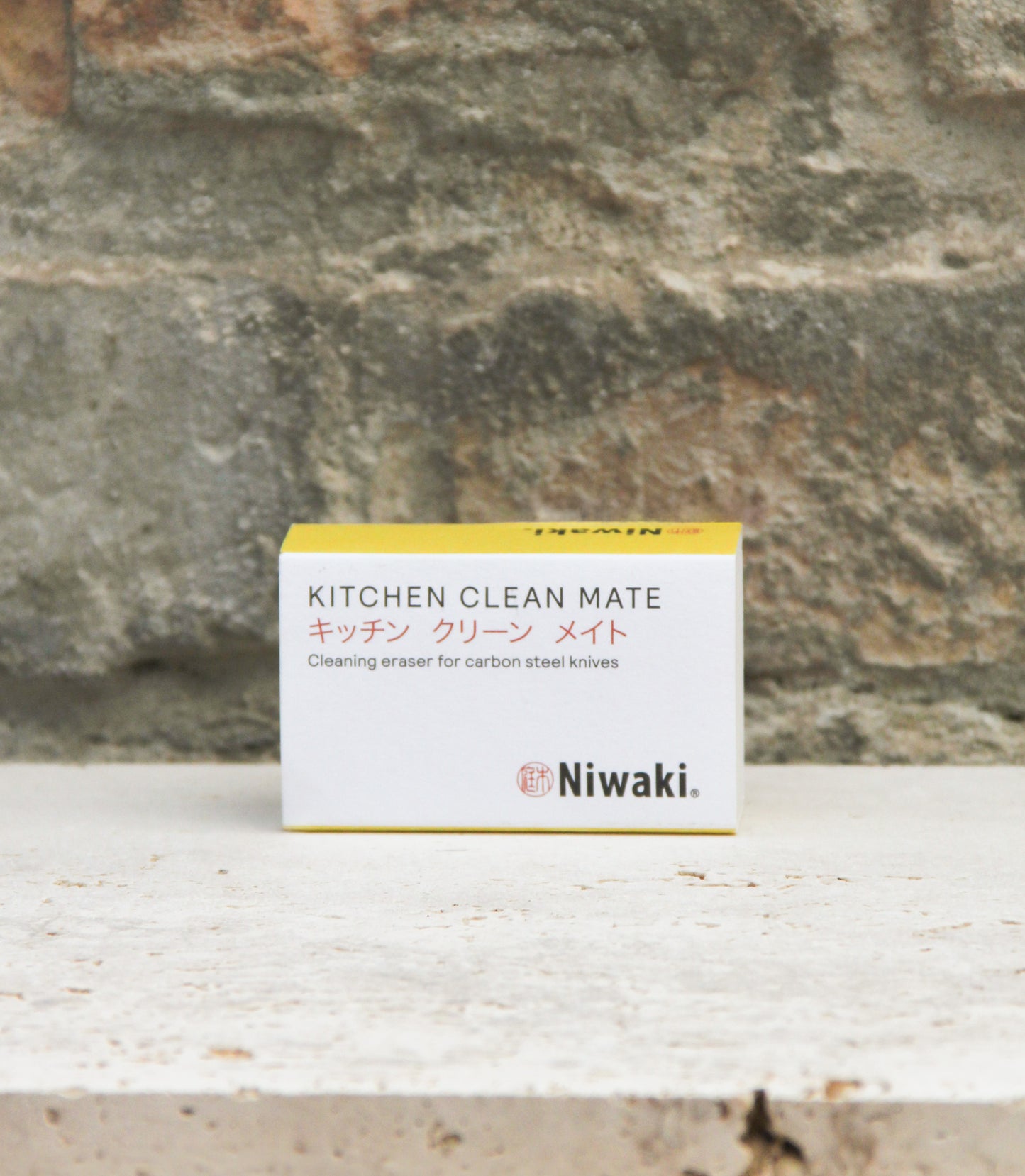 Auf einer hellen Steinoberfläche, vor dem Hintergrund einer strukturierten Ziegelwand, steht eine kompakte Schachtel mit der Aufschrift „Niwaki | ‚Kitchen Clean Mate‘ Rostentferner für Küchenmesser von Niwaki“. Die weiße Schachtel mit gelben Akzenten enthält einen Reinigungsradierer, der speziell für Küchenmesser aus Kohlenstoffstahl entwickelt wurde.