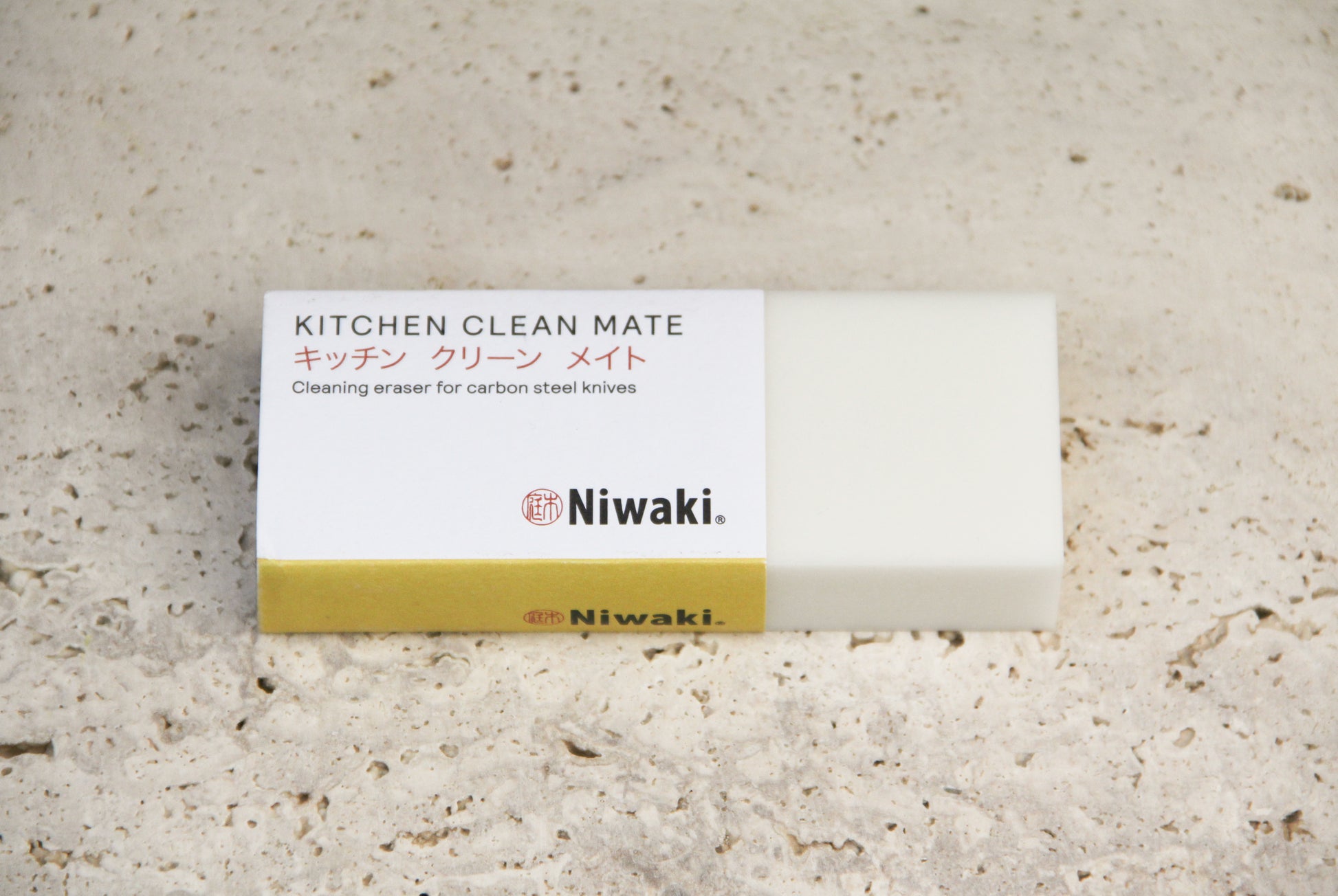 Ein rechteckiger, weiß-goldfarbener Radiergummi mit der Aufschrift „Niwaki | ‚Kitchen Clean Mate‘ Rostentferner für Küchenmesser von Niwaki“, ideal zum Reinigen von Messern aus Kohlenstoffstahl, liegt auf einer strukturierten, beigen Oberfläche.