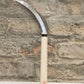 Die Niwaki | „Herbaceous Sickle“ Nokogama Reissichel aus Kohlenstoffstahl, 32 cm – mit ihrer elegant geschwungenen Klinge aus Kohlenstoffstahl und dem Holzgriff – steht aufrecht vor dem Hintergrund einer strukturierten Ziegelwand und verkörpert ein unverzichtbares Gartenwerkzeug, das von der traditionellen japanischen Sichel inspiriert ist.