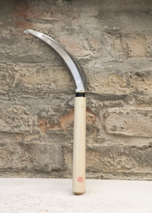 Die Niwaki | „Herbaceous Sickle“ Nokogama Reissichel aus Kohlenstoffstahl, 32 cm – mit ihrer elegant geschwungenen Klinge aus Kohlenstoffstahl und dem Holzgriff – steht aufrecht vor dem Hintergrund einer strukturierten Ziegelwand und verkörpert ein unverzichtbares Gartenwerkzeug, das von der traditionellen japanischen Sichel inspiriert ist.