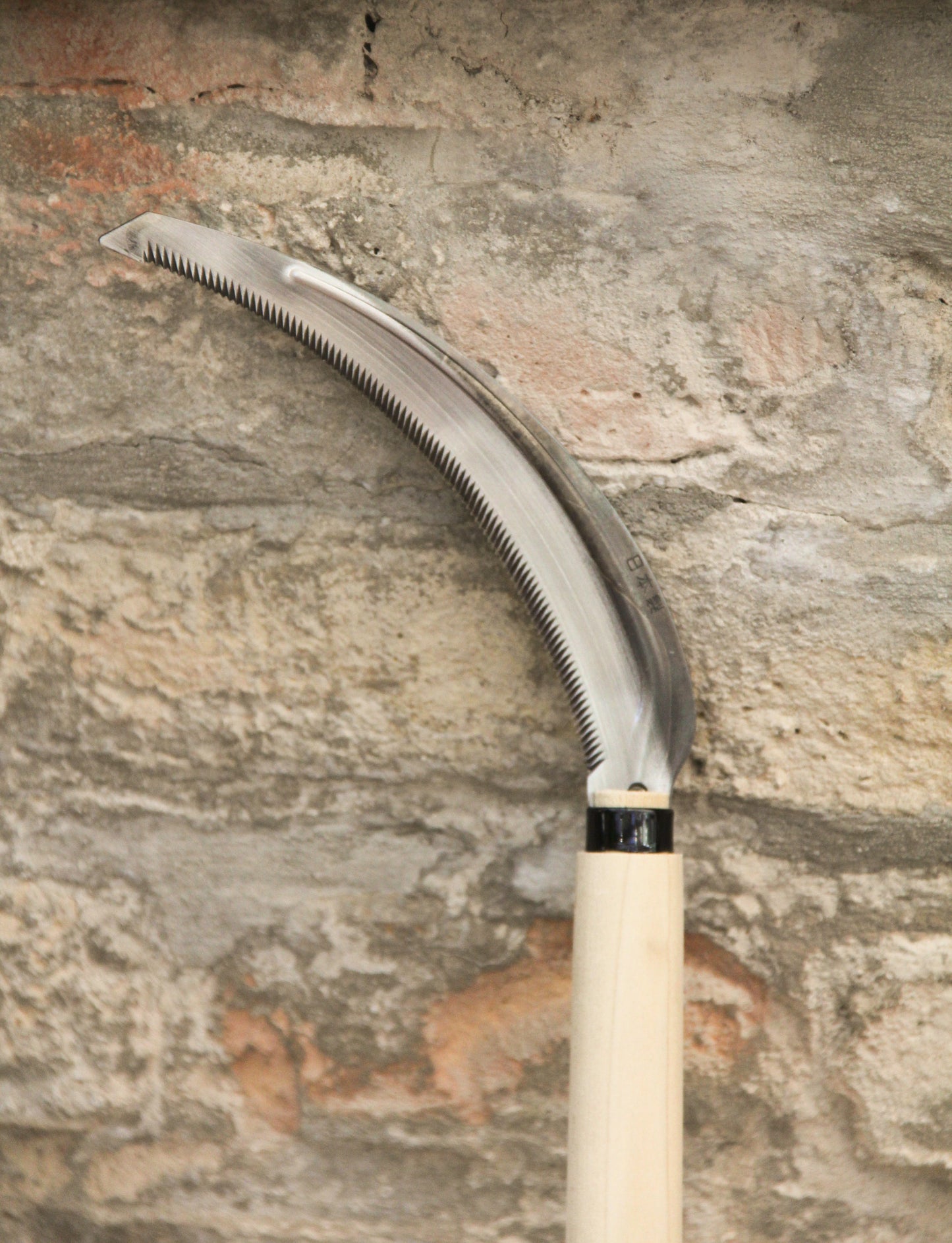 Eine Niwaki „Herbaceous Sickle“ Nokogama Reissichel aus Kohlenstoffstahl (32 cm) mit gezahnter Klinge und einem leichten Holzgriff lehnt an einer strukturierten Steinmauer – ein unverzichtbares Gartenwerkzeug von Niwaki für präzisen Schnitt.