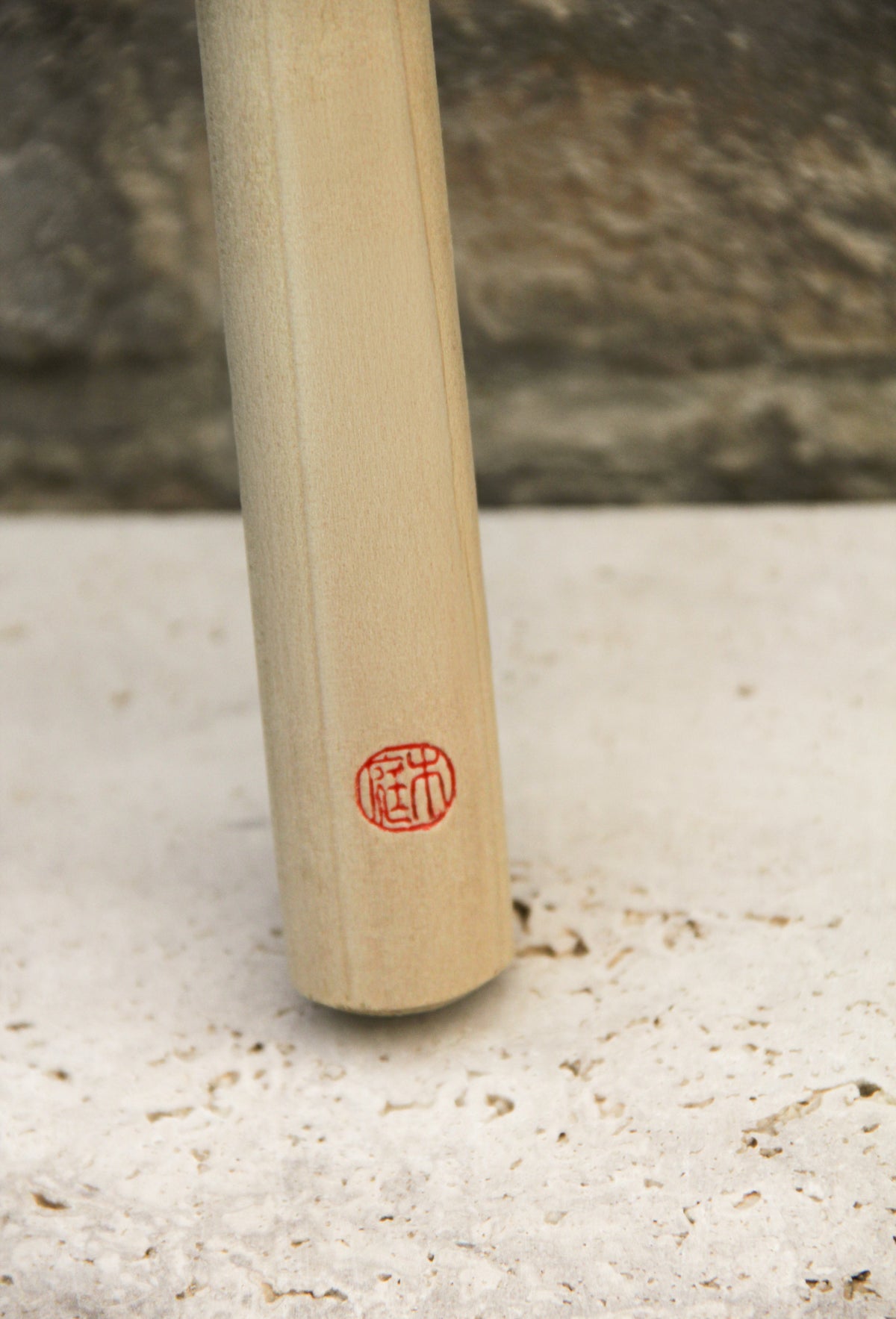 Eine Niwaki „Herbaceous Sickle“ Nokogama Reissichel aus Kohlenstoffstahl (32 cm), die sich durch ihren Holzgriff und einen roten, runden Stempel mit asiatischen Schriftzeichen auszeichnet, ruht auf einer strukturierten, hellen Steinoberfläche, im Hintergrund ist eine verschwommene Steinmauer zu sehen.