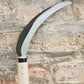 Eine Nahaufnahme einer Niwaki | „Herbaceous Sickle“ Nokogama Reissichel aus Kohlenstoffstahl, 32 cm – mit ihrer glänzenden, geschwungenen Klinge und dem hellen Holzgriff –, die an einer strukturierten Steinmauer lehnt.