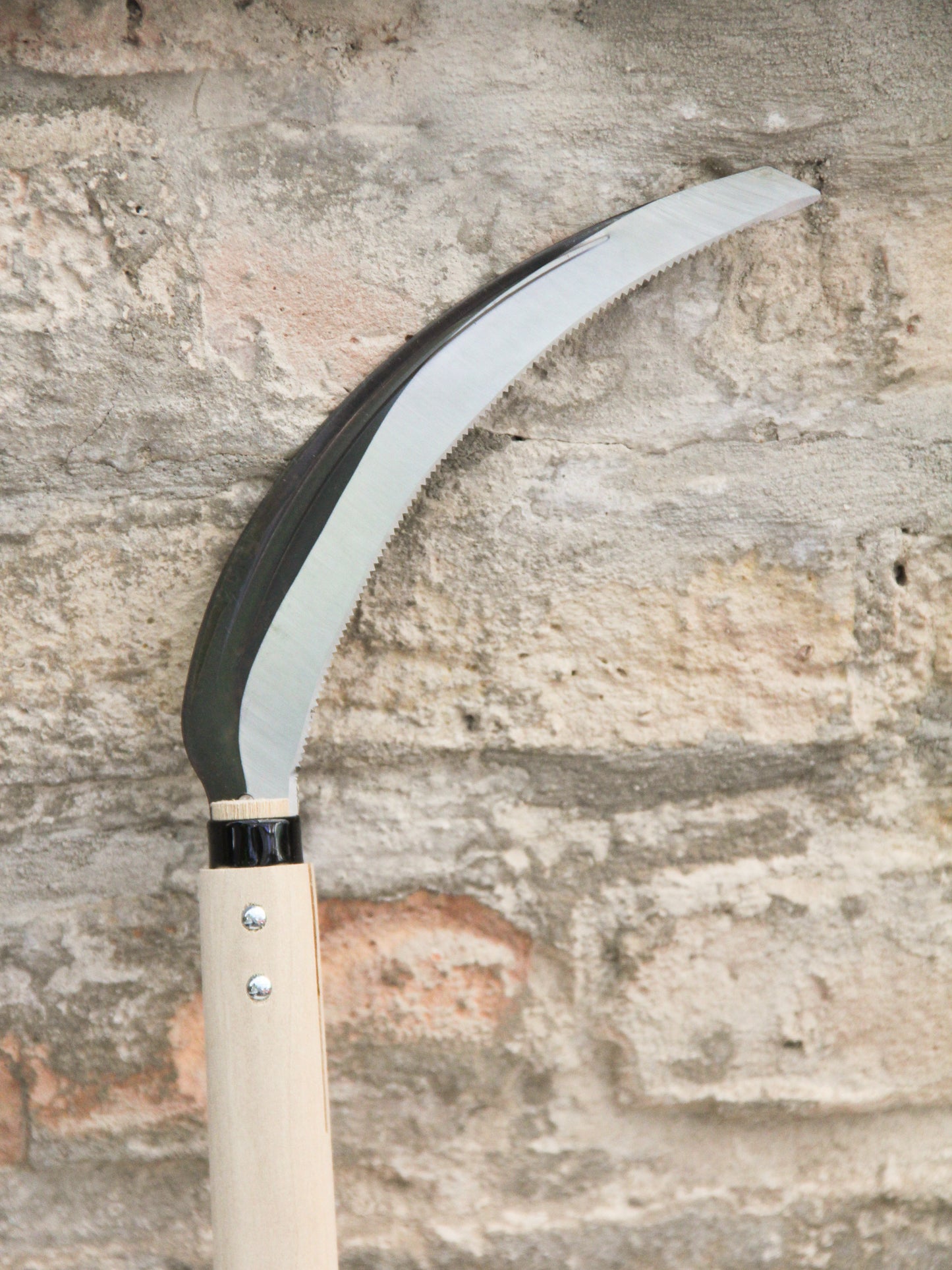 Eine Nahaufnahme einer Niwaki | „Herbaceous Sickle“ Nokogama Reissichel aus Kohlenstoffstahl, 32 cm – mit ihrer glänzenden, geschwungenen Klinge und dem hellen Holzgriff –, die an einer strukturierten Steinmauer lehnt.