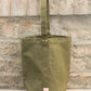 Die „Niwaki | Mini Kantan Werkzeugtasche grün, 28 cm hoch“ von Niwaki besteht aus wasserabweisendem Canvas, ist mit einem einzigen Tragegriff ausgestattet und wird hier aufrecht an einer rustikalen, strukturierten Steinmauer abgebildet. Diese Gartengerätetasche zeichnet sich durch sichtbare Nähte, strapazierfähige Metallnieten und einen kleinen Logo-Aufnäher nahe der unteren Naht aus.