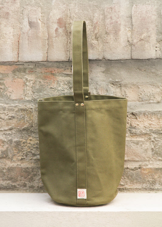 Die „Niwaki | Mini Kantan Werkzeugtasche grün, 28 cm hoch“ von Niwaki besteht aus wasserabweisendem Canvas, ist mit einem einzigen Tragegriff ausgestattet und wird hier aufrecht an einer rustikalen, strukturierten Steinmauer abgebildet. Diese Gartengerätetasche zeichnet sich durch sichtbare Nähte, strapazierfähige Metallnieten und einen kleinen Logo-Aufnäher nahe der unteren Naht aus.