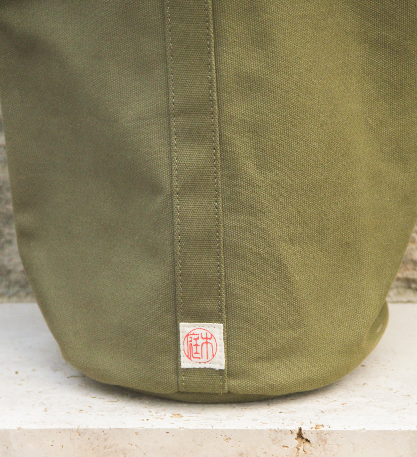 Eine Nahaufnahme der Niwaki | Mini Kantan Werkzeugtasche grün, 28 cm hoch – eine robuste, wasserabweisende Canvas-Tasche von Niwaki in Olivgrün, zeigt die markante vertikale Naht und ein kleines weißes Aufnäher mit roten asiatischen Schriftzeichen am unteren Rand – ideal als Tasche für Gartengeräte.