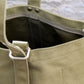 Nahaufnahme der „Niwaki | Mini Kantan Werkzeugtasche grün, 28 cm hoch“ von Niwaki: eine olivgrüne, wasserabweisende Canvas-Tasche mit silbernen Metallbeschlägen und Ziernähten, vor einem unscharfen Steinhintergrund.