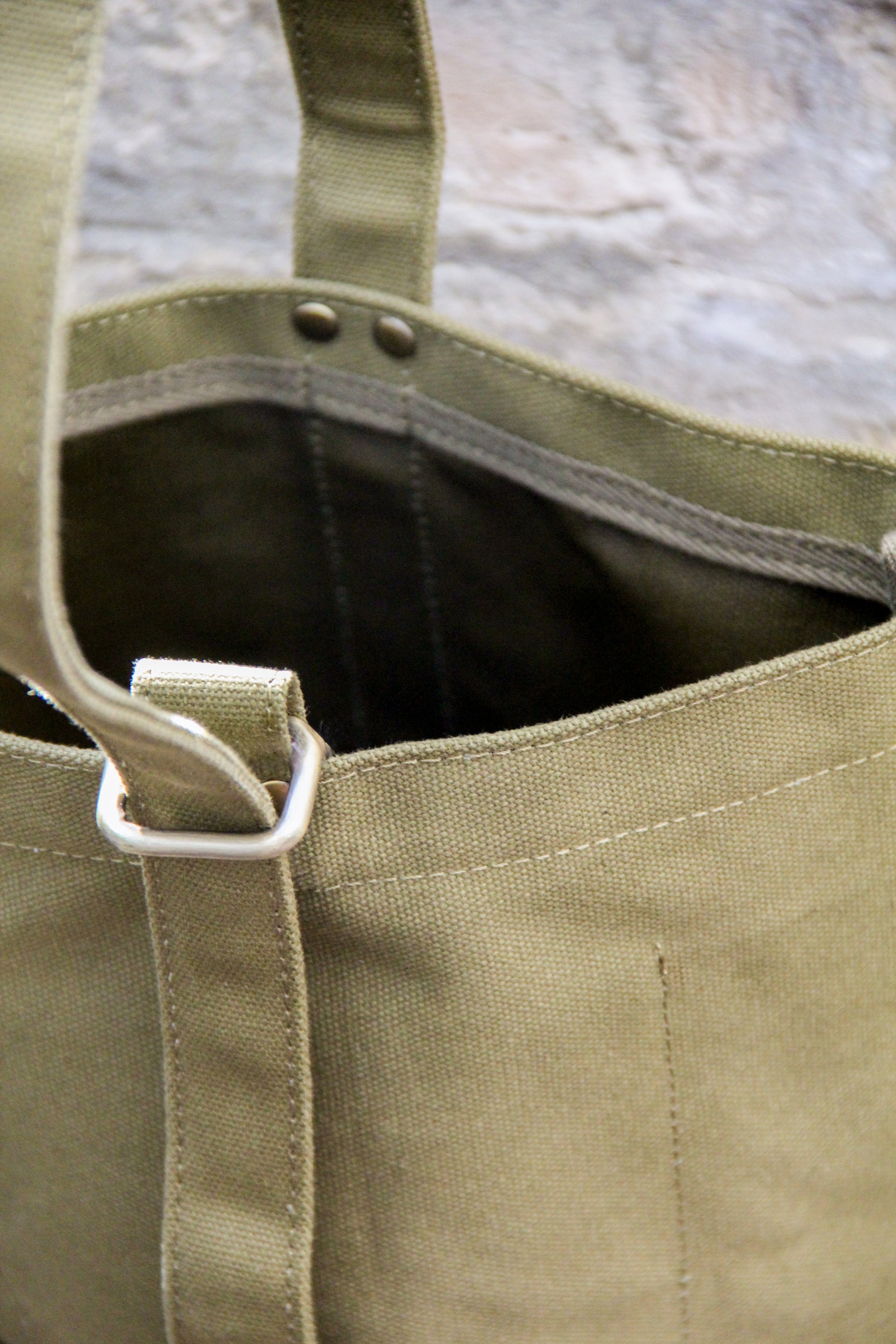 Nahaufnahme der „Niwaki | Mini Kantan Werkzeugtasche grün, 28 cm hoch“ von Niwaki: eine olivgrüne, wasserabweisende Canvas-Tasche mit silbernen Metallbeschlägen und Ziernähten, vor einem unscharfen Steinhintergrund.