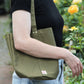 Eine Person in einem schwarzen Hemd und hellblauen Jeans steht neben gelben Rosen und trägt die „Niwaki Mini Kantan Werkzeugtasche“ in Grün (28 cm hoch), eine praktische und wasserabweisende Werkzeugtasche von Niwaki.
