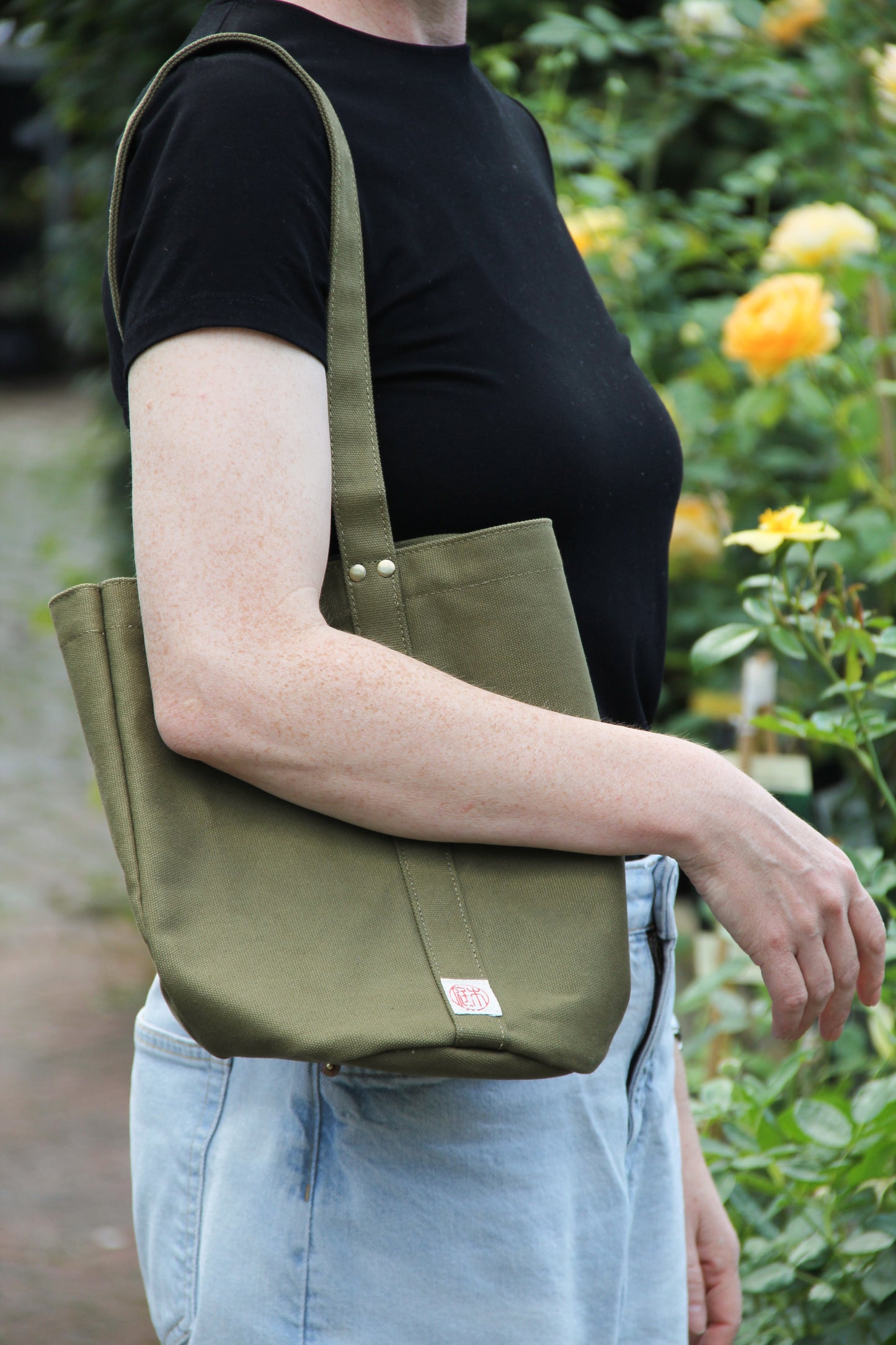 Eine Person in einem schwarzen Hemd und hellblauen Jeans steht neben gelben Rosen und trägt die „Niwaki Mini Kantan Werkzeugtasche“ in Grün (28 cm hoch), eine praktische und wasserabweisende Werkzeugtasche von Niwaki.