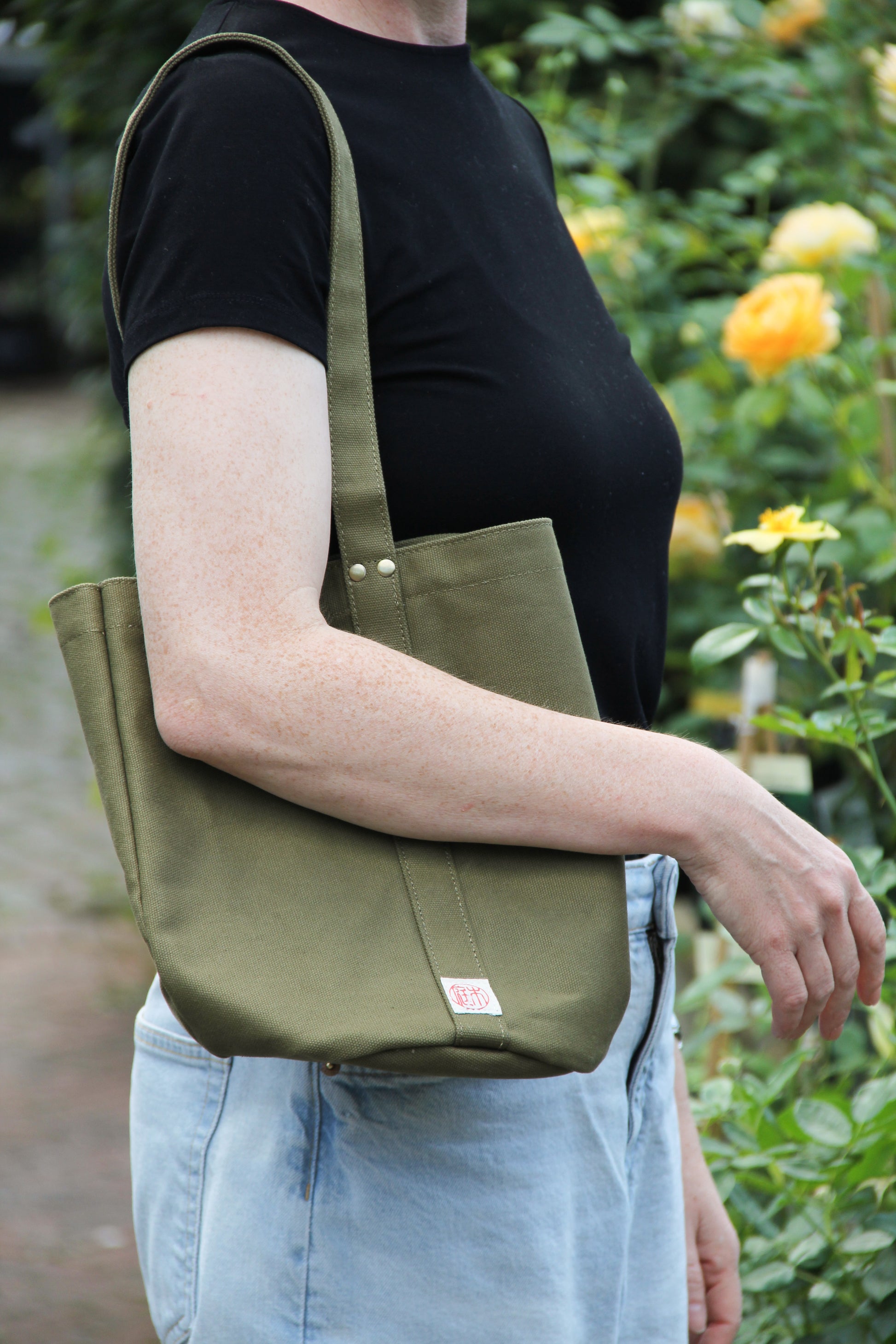 Eine Person in einem schwarzen Hemd und hellblauen Jeans steht neben gelben Rosen und trägt die „Niwaki Mini Kantan Werkzeugtasche“ in Grün (28 cm hoch), eine praktische und wasserabweisende Werkzeugtasche von Niwaki.