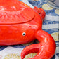 Eine glänzende, leuchtend rote Keramikschale „Summer Bliss Crab“ von Costa Nova mit Deckel und detailgetreuen Scheren ruht auf einem gemusterten Tuch, das mit blauen Meeresmotiven und gelben Kreisen verziert ist, und unterstreicht so den lebendigen Charme der portugiesischen Keramik aus Costa Nova.