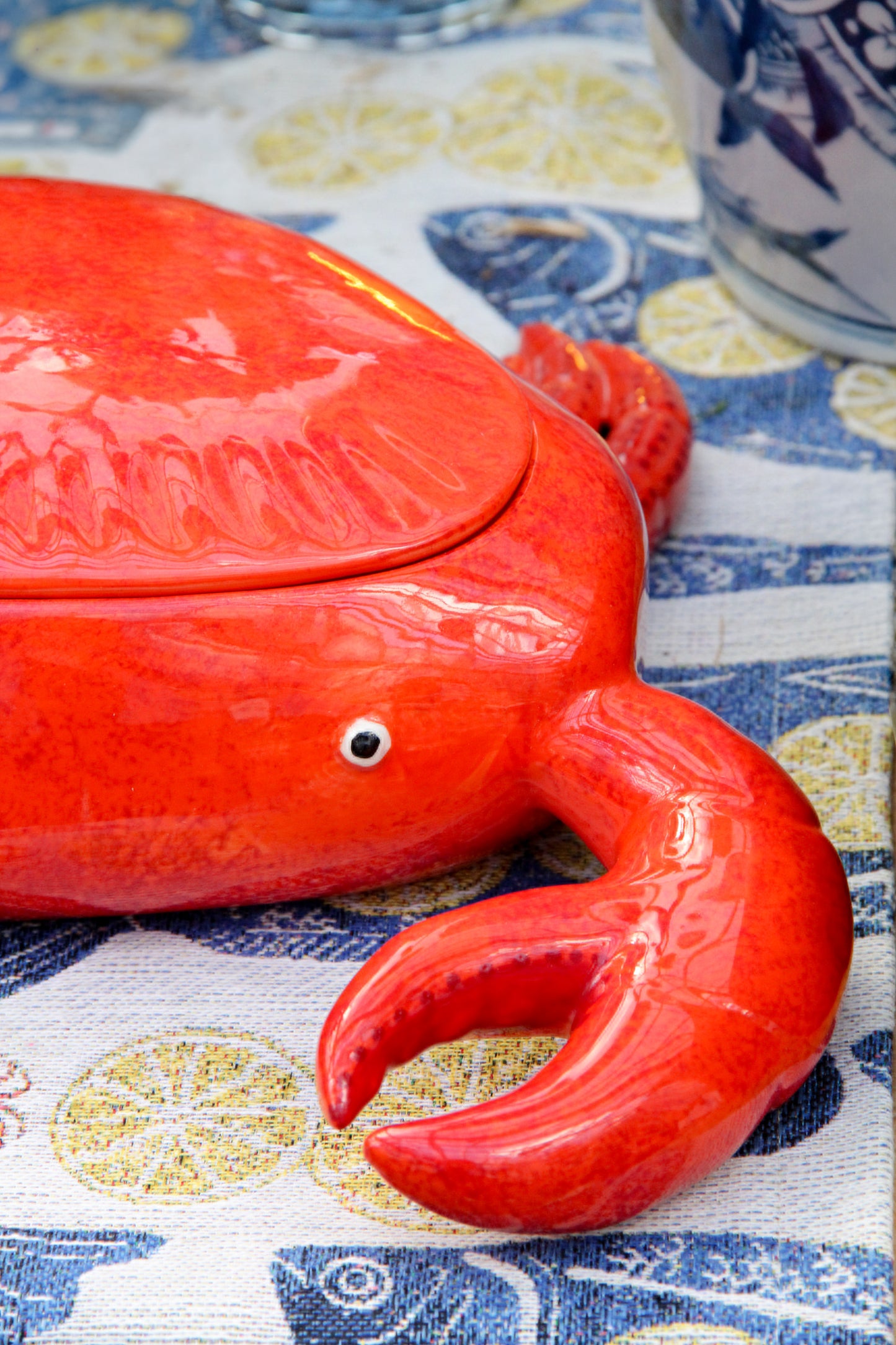 Eine glänzende, leuchtend rote Keramikschale „Summer Bliss Crab“ von Costa Nova mit Deckel und detailgetreuen Scheren ruht auf einem gemusterten Tuch, das mit blauen Meeresmotiven und gelben Kreisen verziert ist, und unterstreicht so den lebendigen Charme der portugiesischen Keramik aus Costa Nova.