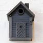 Ein stilvolles schwarzes Vogelhäuschen aus Holz, das „Nistkasten & Futterhaus in einem stilvollen Design | Carbon Black“ von Wildlife Garden, mit einem runden Eingang, steht vor einer weißen Backsteinmauer. An seiner Vorderseite befindet sich unterhalb des Eingangslochs eine kleine rechteckige Tür mit Chevron-Muster, was es zu einem idealen Futterspender für Wildvögel macht.