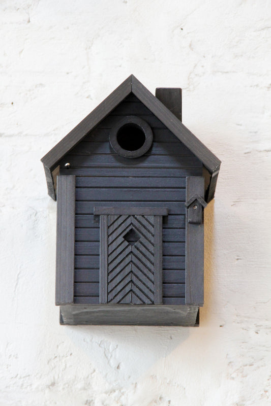 Ein stilvolles schwarzes Vogelhäuschen aus Holz, das „Nistkasten & Futterhaus in einem stilvollen Design | Carbon Black“ von Wildlife Garden, mit einem runden Eingang, steht vor einer weißen Backsteinmauer. An seiner Vorderseite befindet sich unterhalb des Eingangslochs eine kleine rechteckige Tür mit Chevron-Muster, was es zu einem idealen Futterspender für Wildvögel macht.