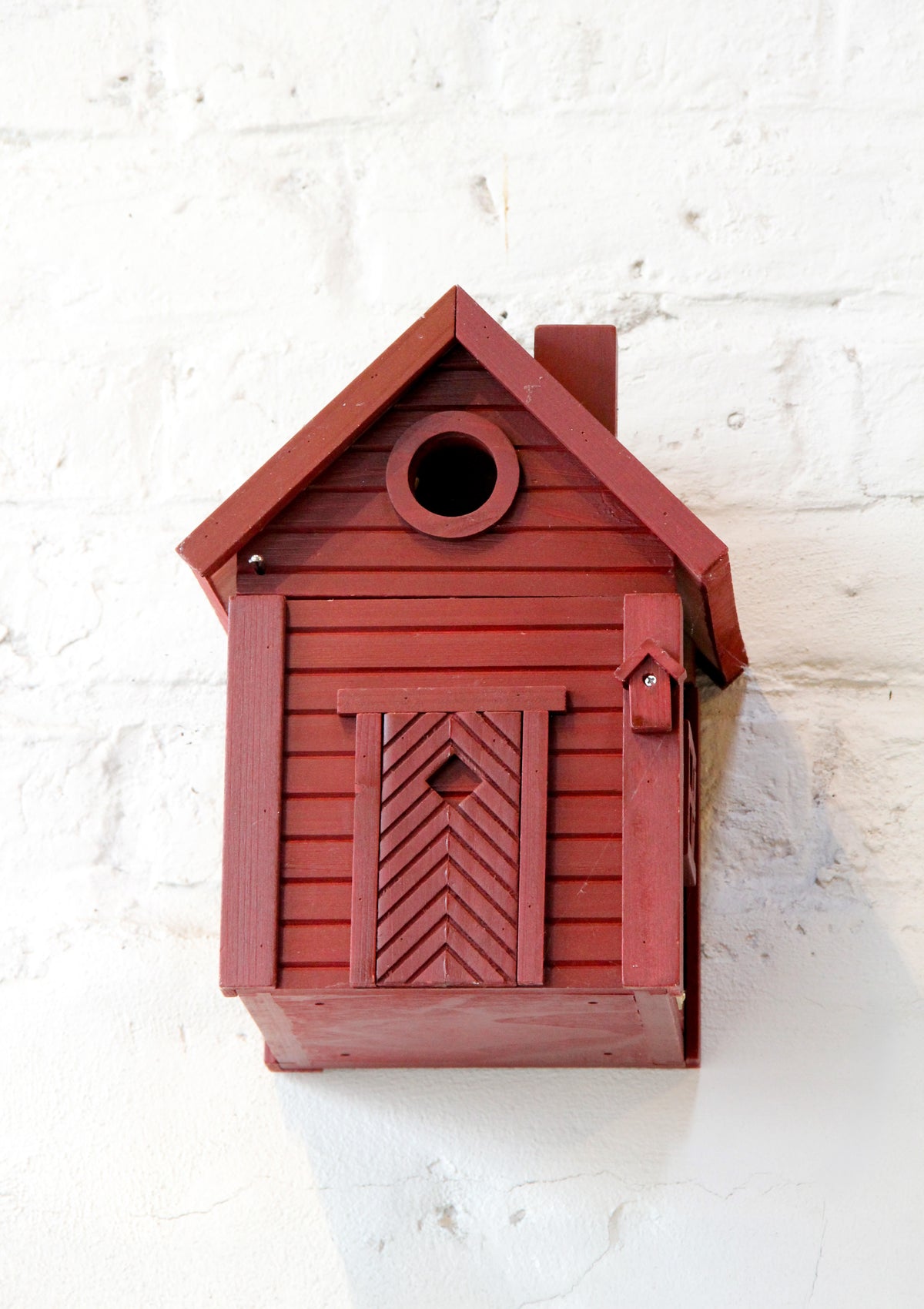 Ein stilvolles „Nistkasten & Futterhaus in einem stilvollen Design | Red Earth“ von Wildlife Garden, bestehend aus einer charmanten roten Holzkonstruktion mit schrägem Dach und einer kleinen Ziertür mit herzförmigem Ausschnitt, ist an einer weißen Ziegelwand angebracht. Dank seiner runden Einflugöffnung ist dieser Nistkasten sowohl funktional als auch ein echter Blickfang für Ihre gefiederten Freunde.