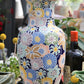 Eine bunte „Des Pots“-Vase aus glasierter Keramik mit Blumenmuster (H 40 cm, T 19,5 cm) steht auf einer Holzunterlage, umgeben von verschiedenen Pflanzen, einer Flasche Olivenöl und weiteren dekorativen Elementen in einem hellen Innenraum.