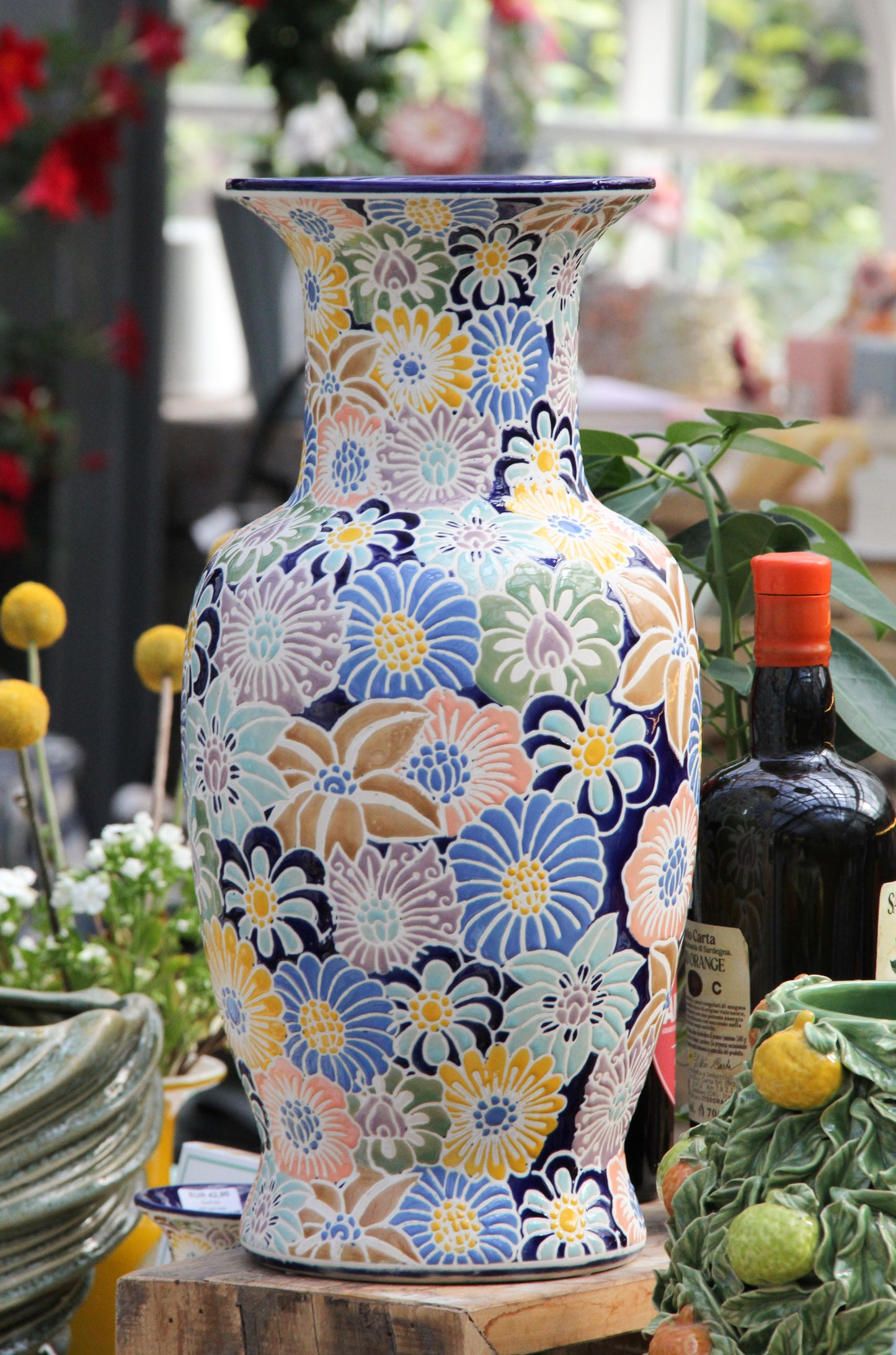 Eine bunte „Des Pots“-Vase aus glasierter Keramik mit Blumenmuster (H 40 cm, T 19,5 cm) steht auf einer Holzunterlage, umgeben von verschiedenen Pflanzen, einer Flasche Olivenöl und weiteren dekorativen Elementen in einem hellen Innenraum.